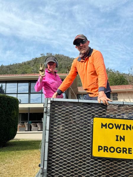 Two workers standing behind a caged trailer that carries a Mowing In Progress sign, on a lawn in front of a large building