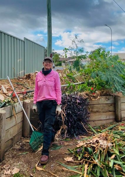 A worker in a pink high visibility shirt standing with a rake beside a large pile of cleared garden waste