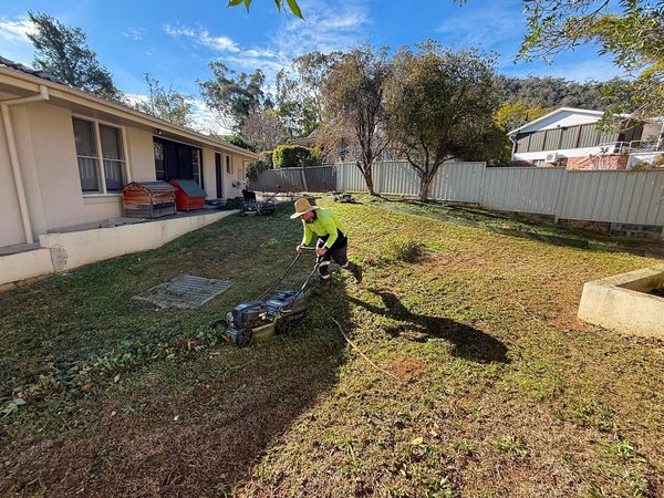 A worker in a lime high visibility shirt and a wide-brimmed hat pushing a mower across a large back lawn
