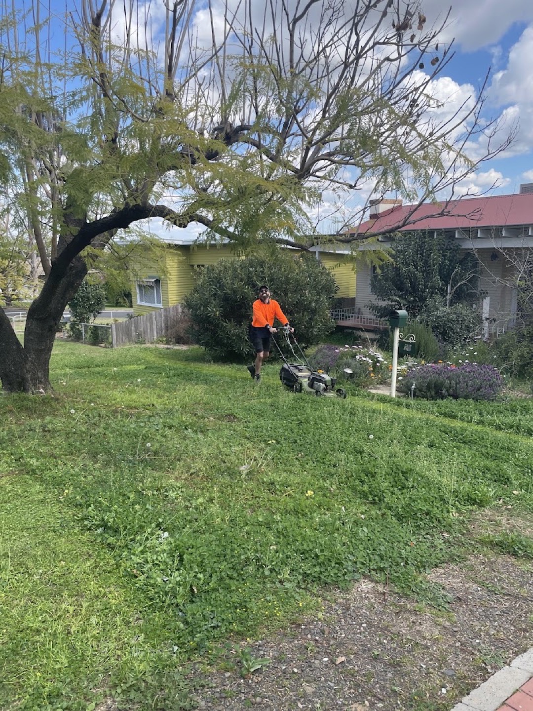 A worker in high visibility orange trimming grass in a back yard beside a brick house