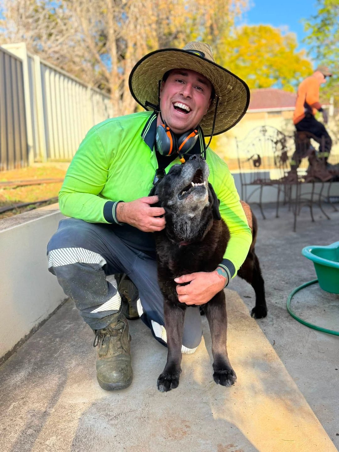 A worker in a lime high visibility shirt and a wide-brimmed hat crouching beside a dog on a paved area, with a second worker cutting back a garden behind