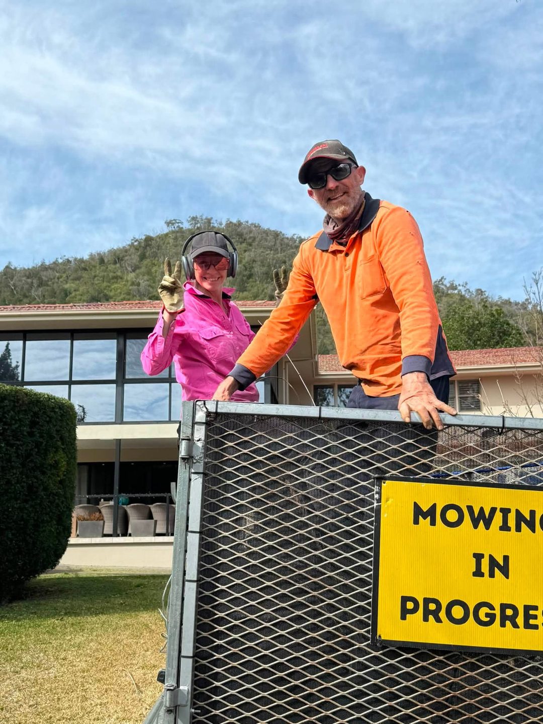 Two workers standing behind a caged trailer that carries a Mowing In Progress sign, on a lawn in front of a large building