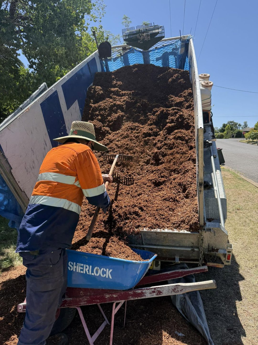 A worker in orange high visibility gear raking mulch off the back of a tipping truck into a wheelbarrow