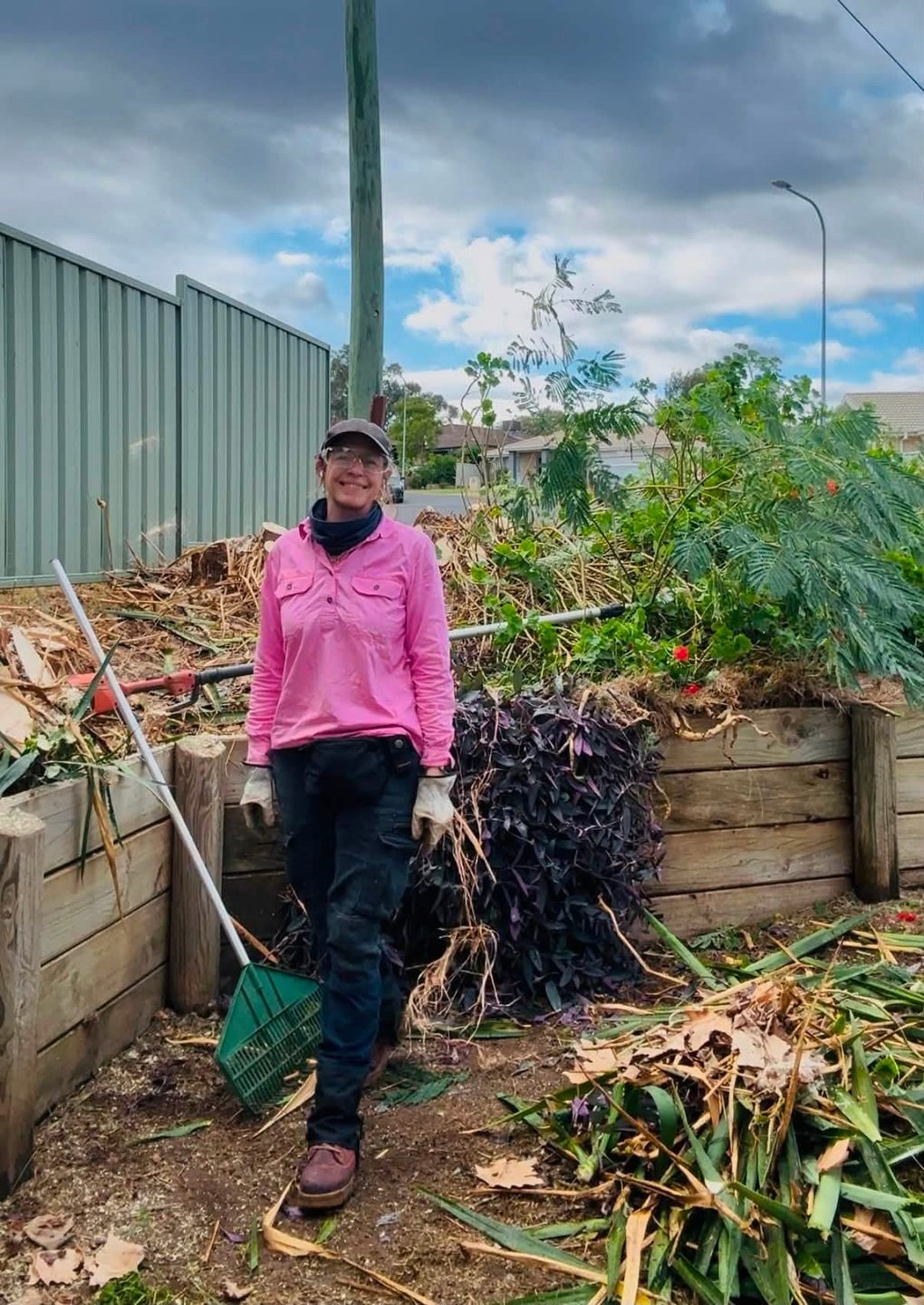 A worker in a pink high visibility shirt standing with a rake beside a large pile of cleared garden waste